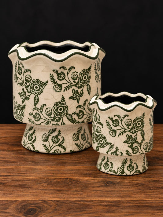 Dos maceteros de cerámica craquelada con flores verde oliva y borde ondulado, grande y pequeño, sobre mesa de madera